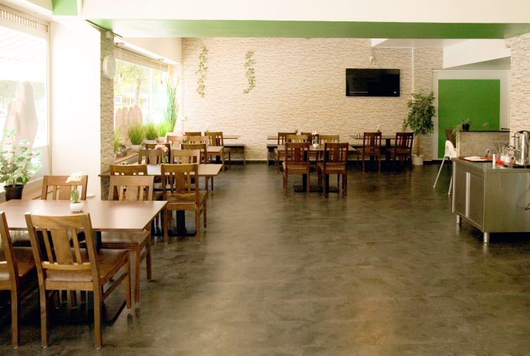 Heller Restaurantbereich mit Holztischen, grünen Pflanzen, Steinwänden und einem Kaffeeautomaten im Hintergrund.