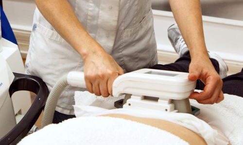 Fachkraft in weißer Uniform lächelt während der Anwendung eines ästhetischen Geräts auf eine liegende Person.