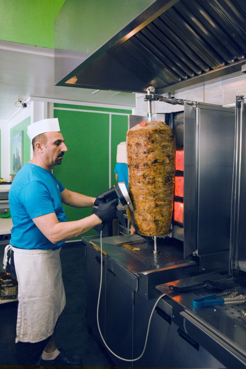 Mann mit weißer Mütze und Schürze schneidet mit einem elektrischen Messer Fleisch von einem vertikalen Grillspieß.