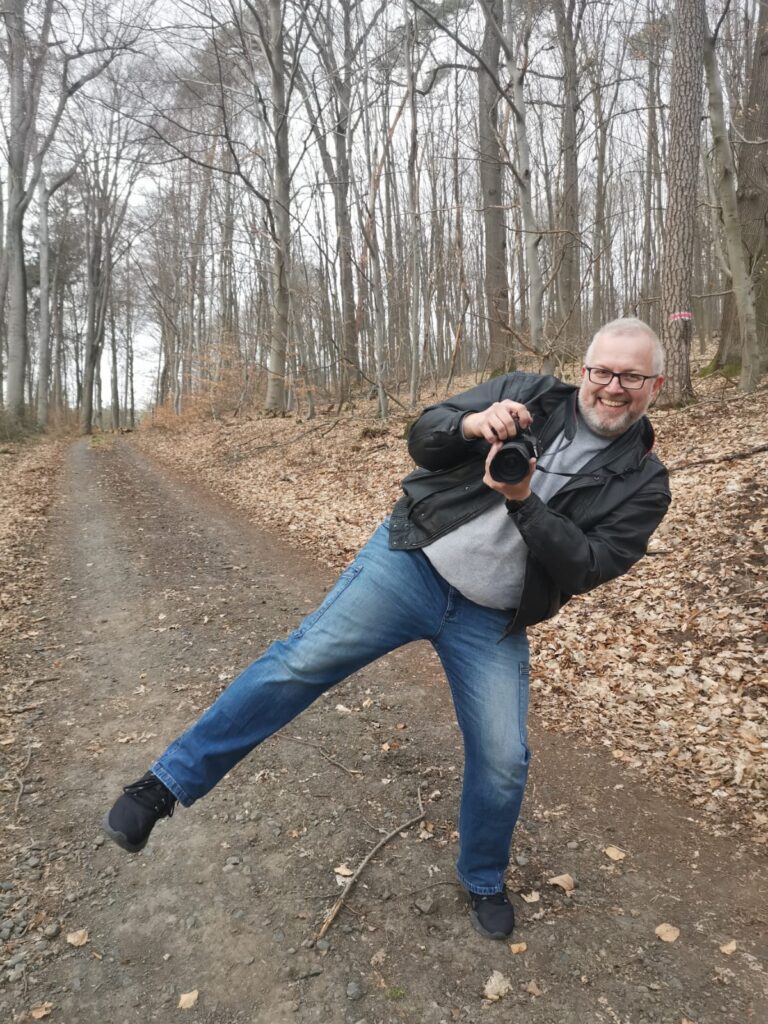Ein fröhlicher Mann mit Kamera posiert spielerisch auf einem Waldweg, hebt ein Bein und lächelt in die Kamera.