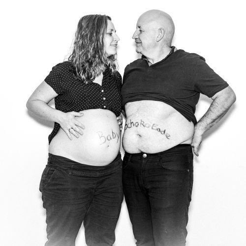 Zwei Erwachsene stehen nebeneinander vor weißem Hintergrund. Beide heben ihre Oberteile und zeigen ihre Bäuche. Auf dem Bauch der linken Person steht ‚Baby‘, auf dem der rechten ‚Schokolade‘. Sie lächeln sich gegenseitig an.