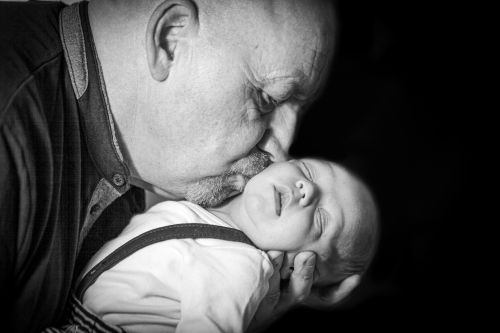Schwarz-weiß-Foto eines älteren Mannes mit Glatze und Schnurrbart, der einem schlafenden Baby zärtlich auf die Wange küsst. Der Mann trägt ein Hemd und hält den Kopf des Babys behutsam mit einer Hand. Das Baby trägt helle Kleidung mit dunklen Hosenträgern.