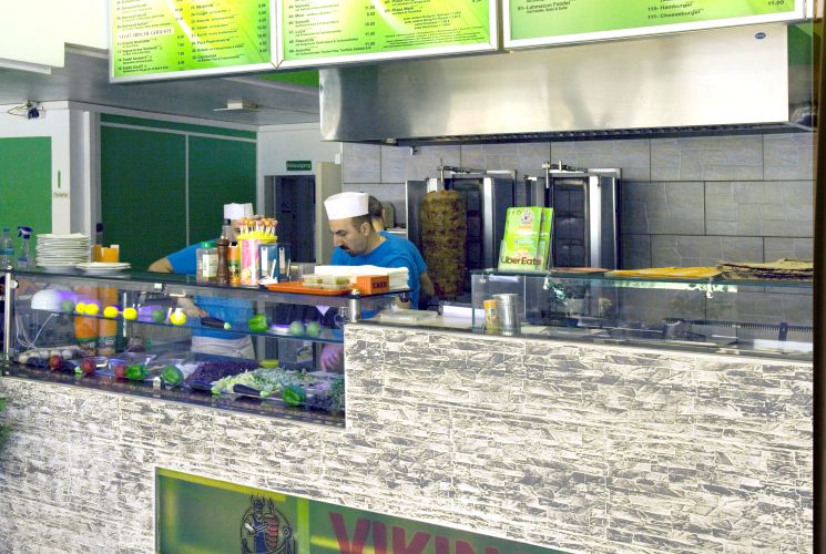 Zwei Mitarbeiter in einem Fast-Food-Restaurant bereiten Speisen hinter einer Theke mit frischen Zutaten und vertikalen Grillspießen zu.