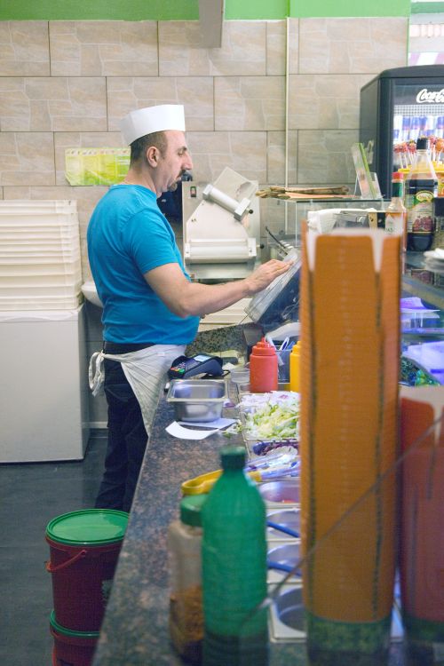 Ein Mann in Arbeitskleidung steht hinter einer Theke mit frischen Zutaten und bedient ein Touchscreen-Gerät in einem Imbiss.