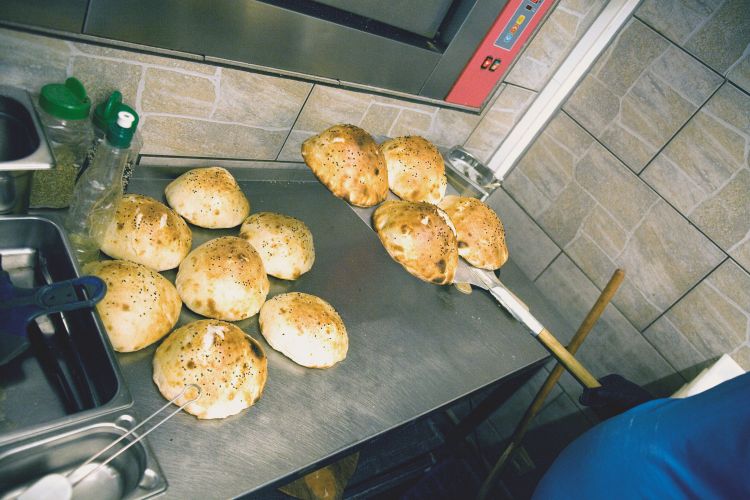 Zehn runde Brote mit Sesam auf einer Metallfläche, ein Mitarbeiter bewegt sie mit einem langen Werkzeug.