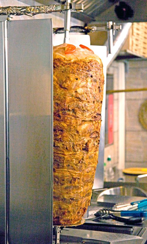 Ein großer, rotierender Fleischspieß mit Tomaten oben wird in einer Metallvorrichtung gegart – typisch für Döner oder Shawarma.
