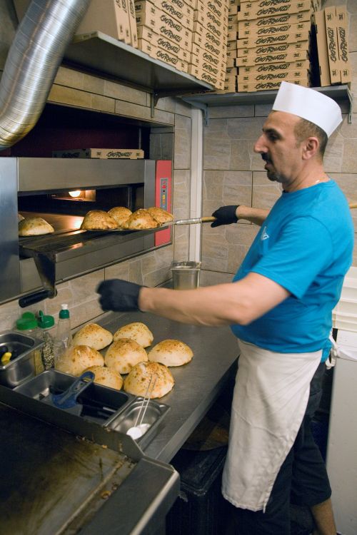 Ein Mann mit weißer Mütze und Schürze holt mit einem langen Metallwerkzeug frisch gebackenes Brot aus einem großen Industrieofen.