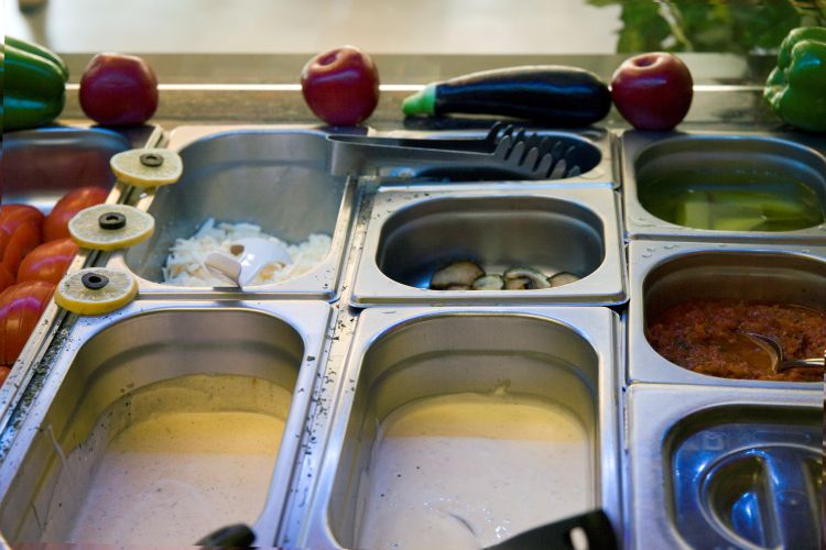 Edelstahlbehälter mit Zutaten wie Tomaten, Käse, Pilzen, Saucen und Oliven in einer Buffetstation mit dekorativem Gemüse
