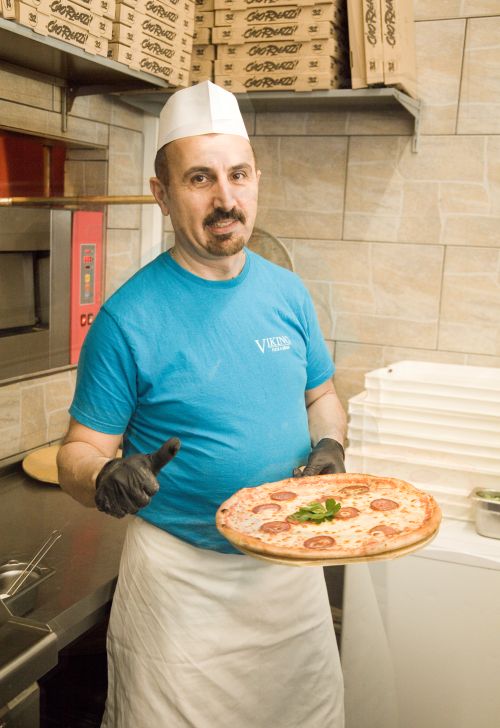 Mann mit Viking-Shirt und Schürze hält eine Pepperoni-Pizza mit Petersilie in einer Küche mit Pizzaofen und Giorgio’s-Kartons.