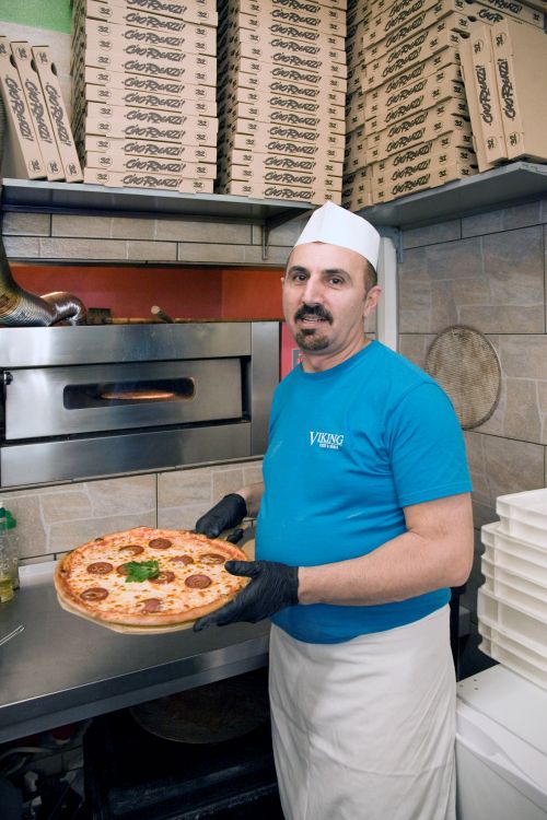 Mann mit Viking Pizza-Shirt und Schürze hält eine frisch gebackene Pepperoni-Pizza in einer Pizzeria-Küche.