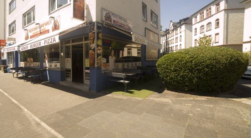 Straßenecke mit Restaurant „Döner Haus“, das Döner, Burger und Pizza anbietet, mit Außenbestuhlung und Werbebildern.
