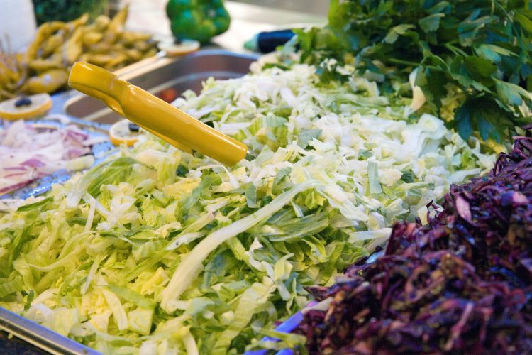 Große Mengen geschnittenes Gemüse wie Eisbergsalat, Rotkohl, Zwiebeln und Petersilie in Edelstahlbehältern mit gelber Zange.