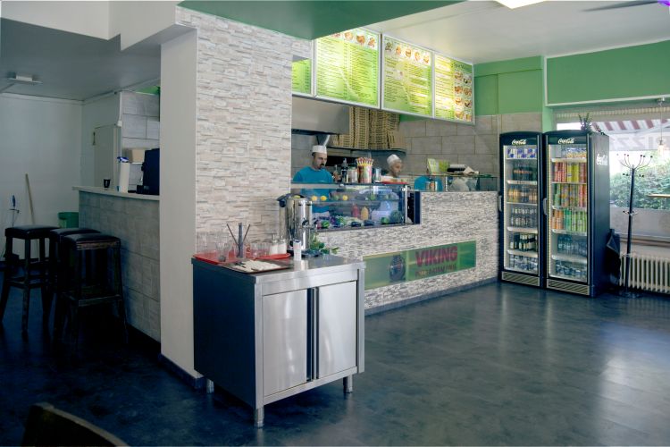 Zwei Mitarbeiter in einem Fast-Food-Restaurant bereiten Speisen hinter einer Theke mit frischen Zutaten und vertikalen Grillspießen zu.